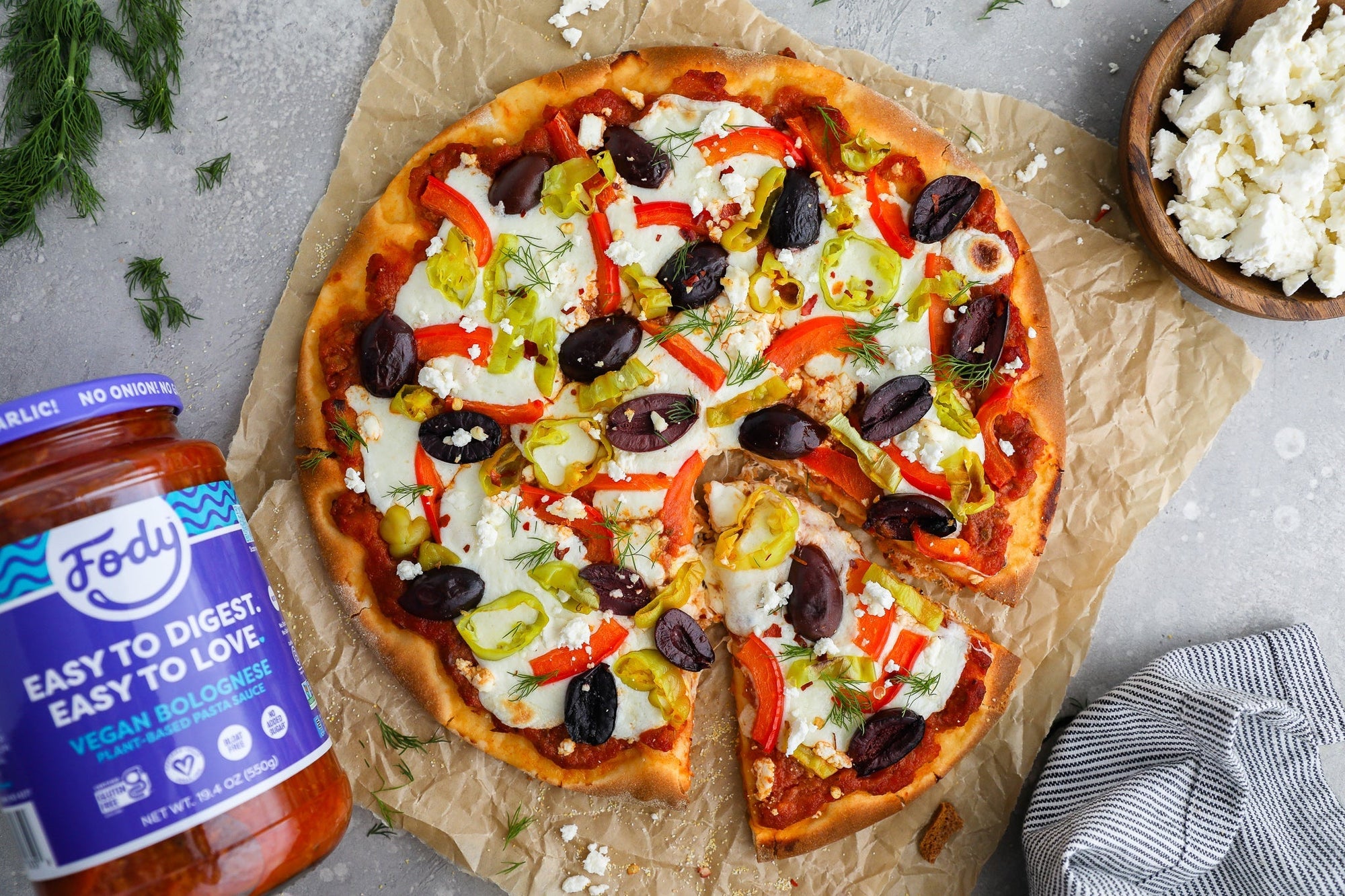 An image of Fody’s gut-friendly Greek pizza. A rustic-looking Greek pizza on a plate beside a bottle of Fody’s Bolognese sauce.