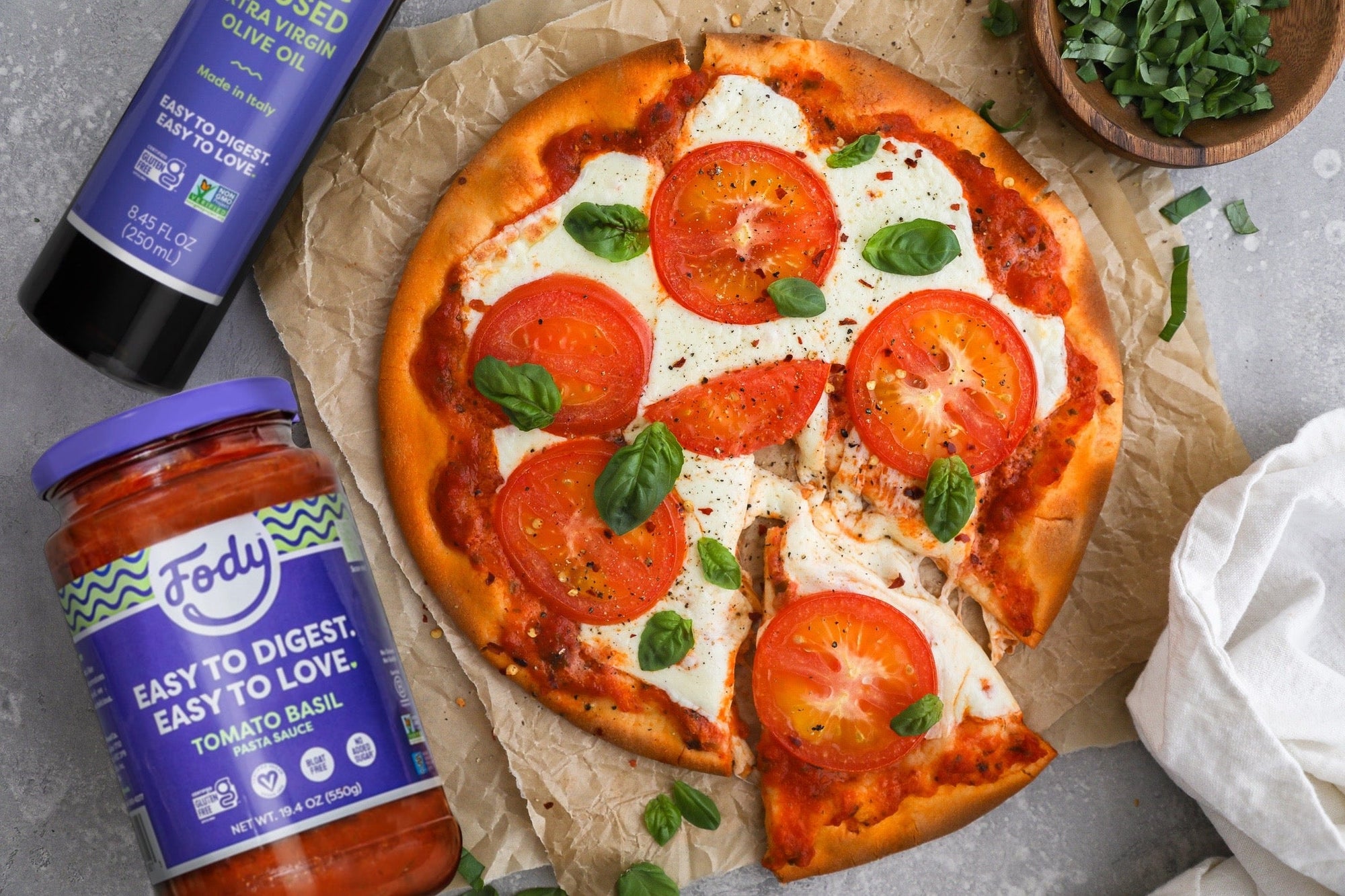 An image of Fody's gluten-free margherita pizza beside two bottles of Fody Tomato Basil sauce and Olive Oil.