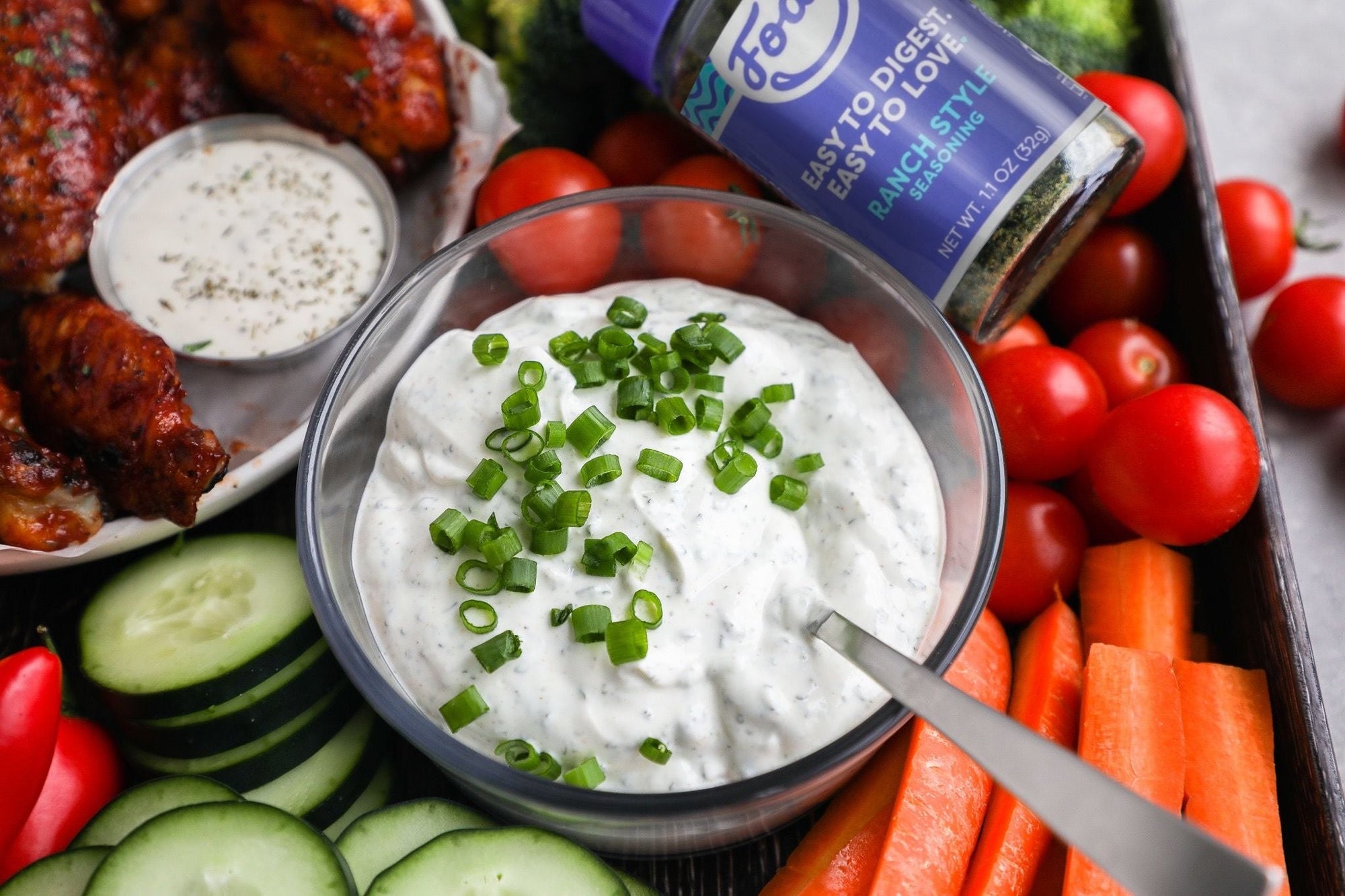 An image of Fody's sliced veggies with homemade ranch dip. Sliced cucumbers, carrots and cherry tomatoes are spread over a plate around a bowl of white homemade ranch dip topped with scallions.