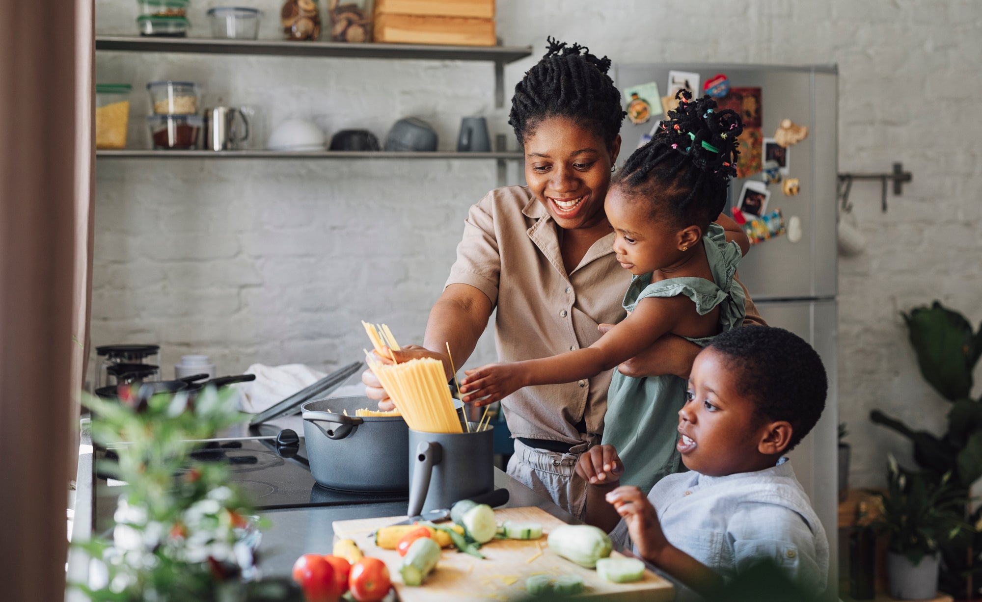 A mother holding her toddler daughter while making a plant-based recipe with her young son.