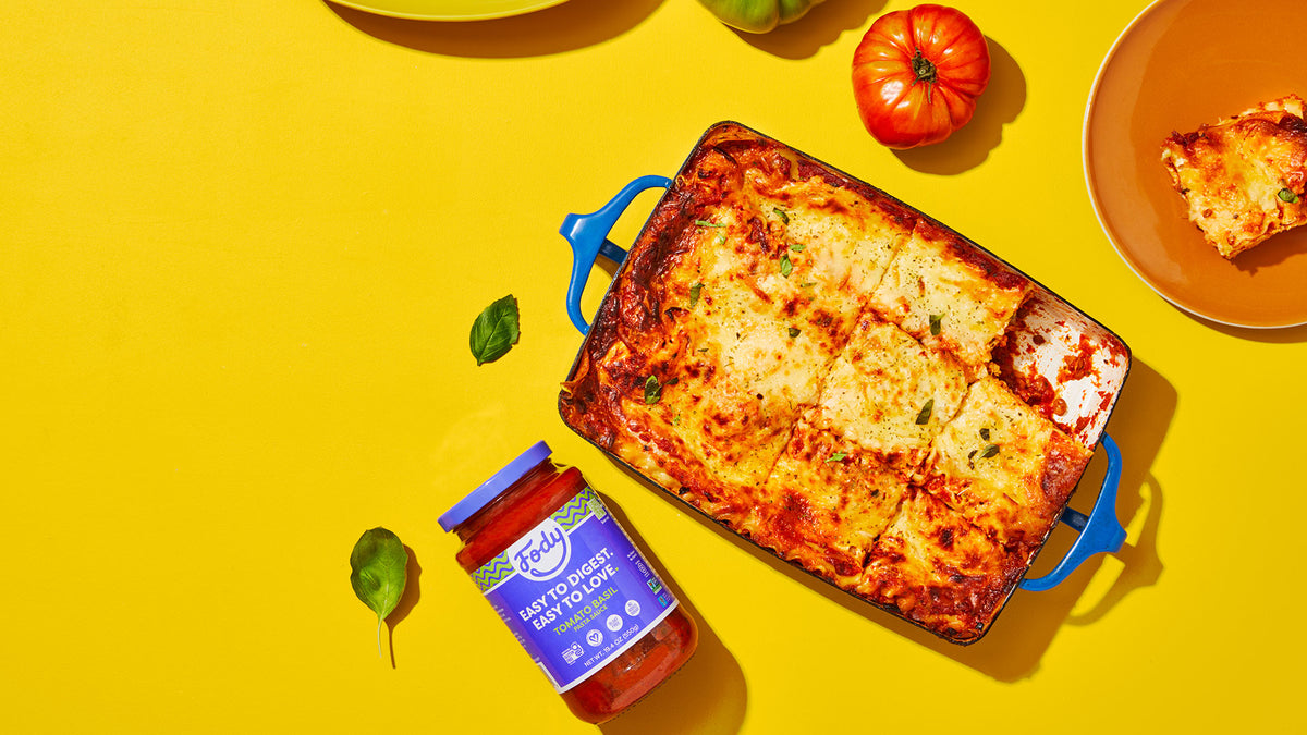 Baked lasagna in a blue dish beside a Fody pasta sauce jar on a yellow background, garnished with basil and tomatoes.