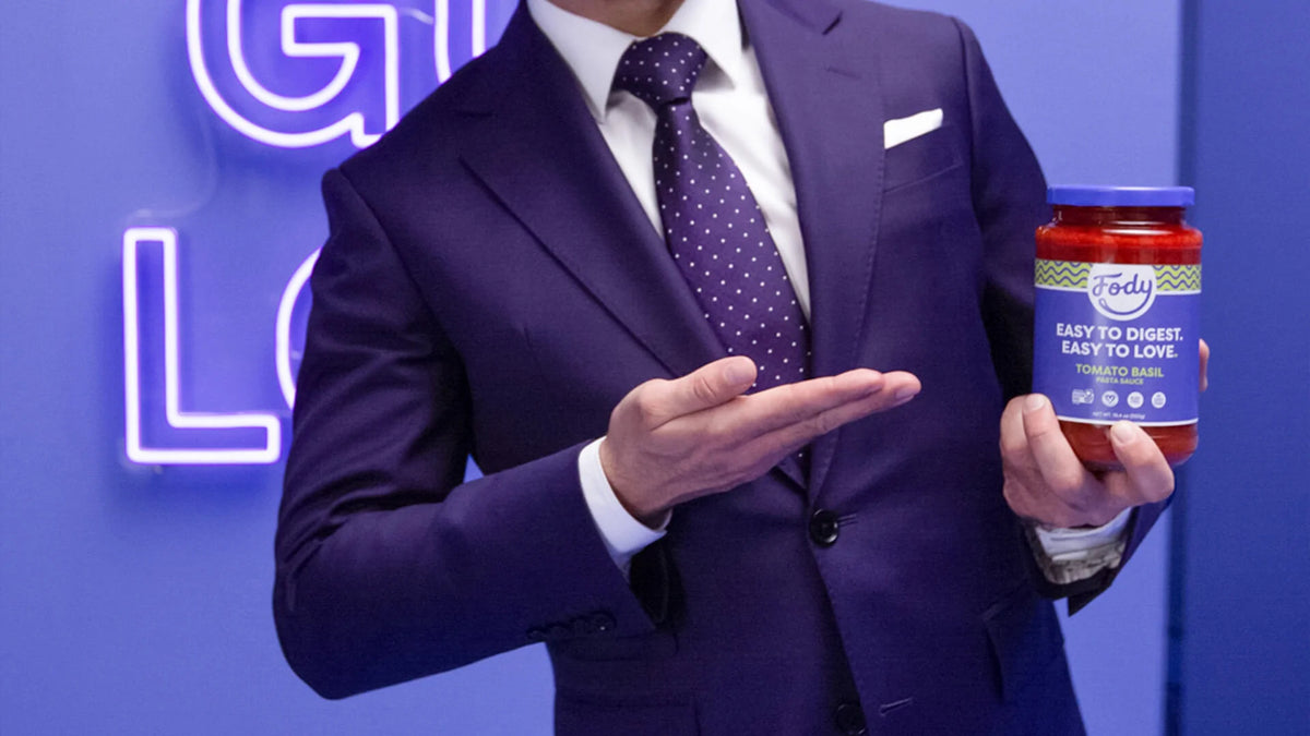 Man in a purple suit pointing to a jar of Fody Tomato Basil Pasta Sauce against a blue background with glowing text.