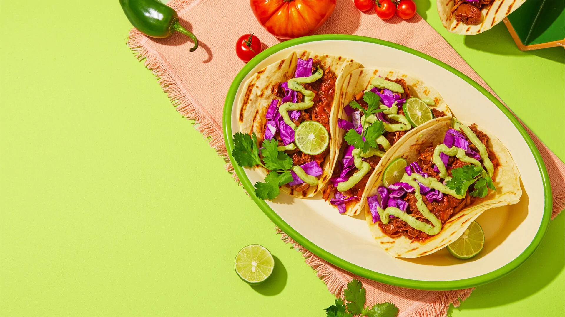 Three tacos filled with seasoned meat, purple cabbage, lime, and cilantro on a green plate against a bright lime background with peppers and tomatoes.