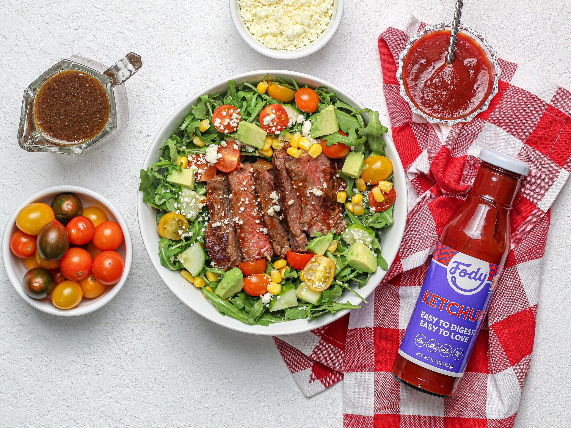 Low FODMAP steak salad with arugula, avocado, corn, and tomatoes beside a Fody ketchup bottle