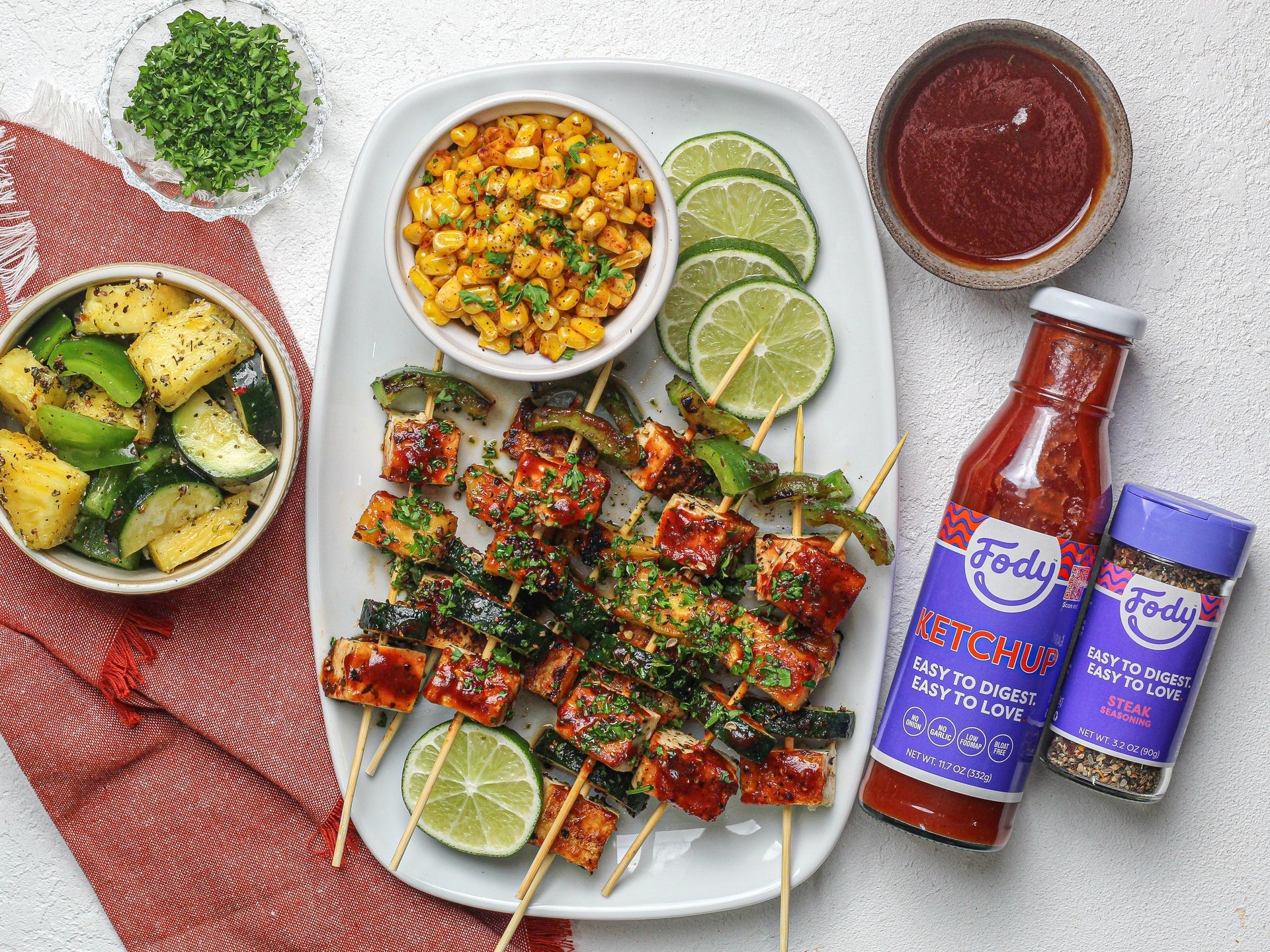 Low FODMAP tofu skewers with grilled zucchini and corn beside a Fody Ketchup bottle and lime wedges