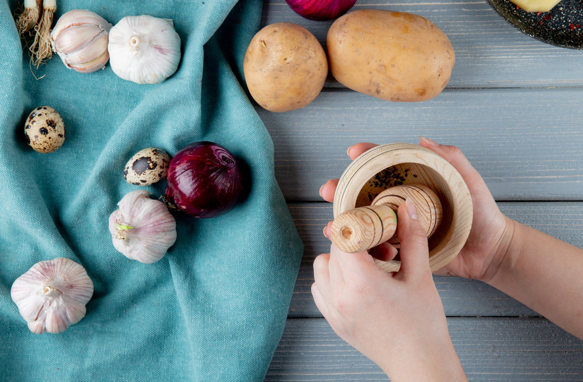 Whole garlic bulbs and a red onion, both alliums to know about with garlic intolerance, beside potatoes as hands grind spices in a wooden mortar