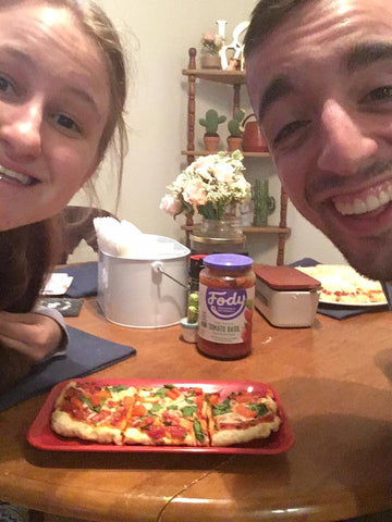 A smiling couple sitting at a table with homemade pizza topped with veggies and a jar of Fody Low FODMAP Tomato Basil Pasta Sauce.