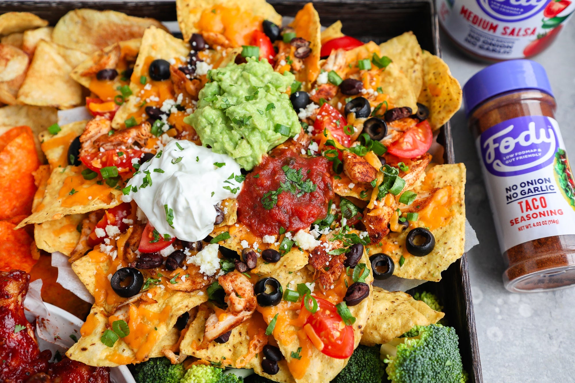 An image of Fody's spicy chicken low FODMAP nachos: yellow nachos topped with vegetables, black olives, guacamole, salsa and sour cream beside a bottle of Fody's taco seasoning.