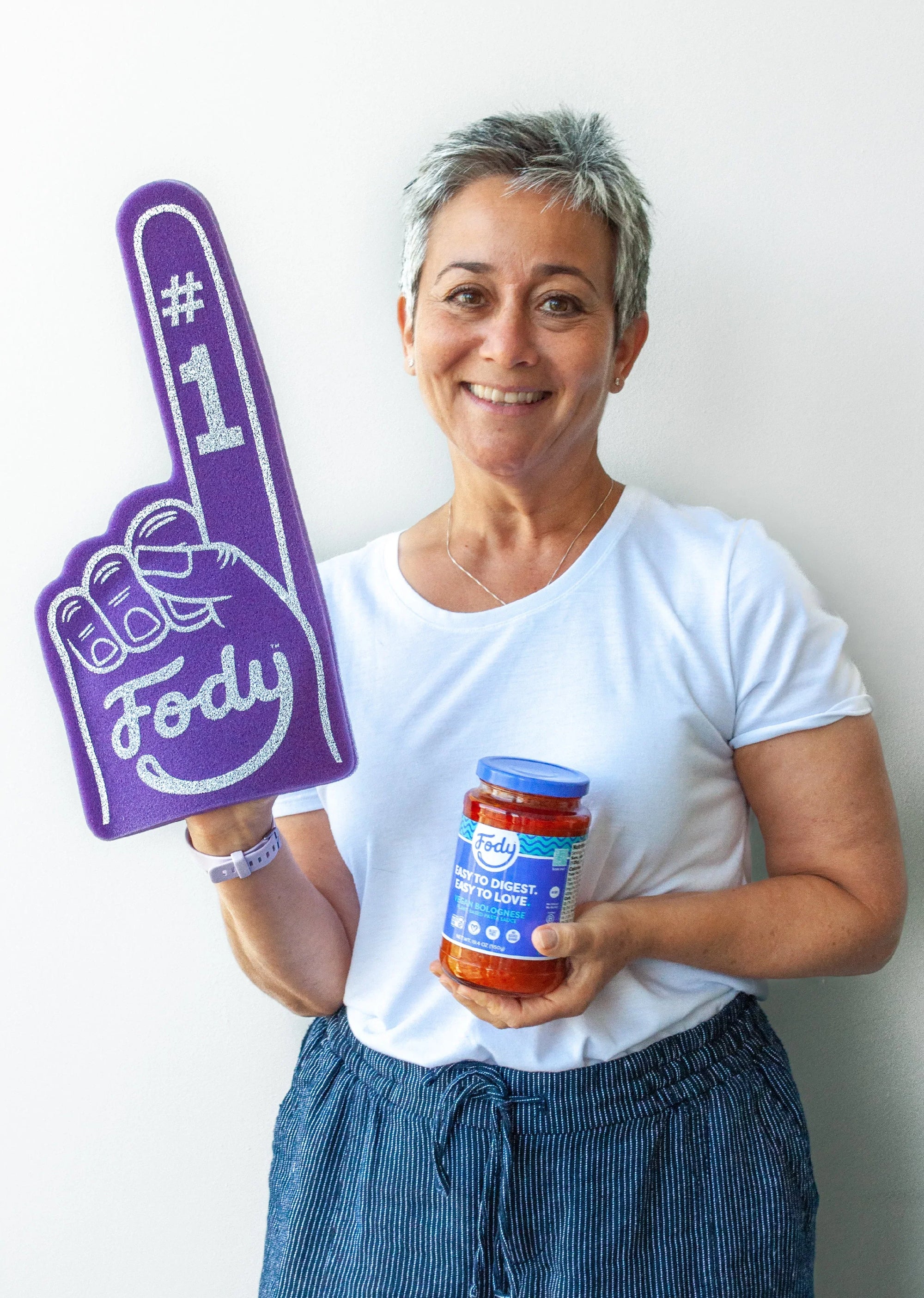 Person smiling while holding a purple “#1 Fody” foam finger and a jar of Fody Bolognese Pasta Sauce, celebrating gut-friendly Low FODMAP food.
