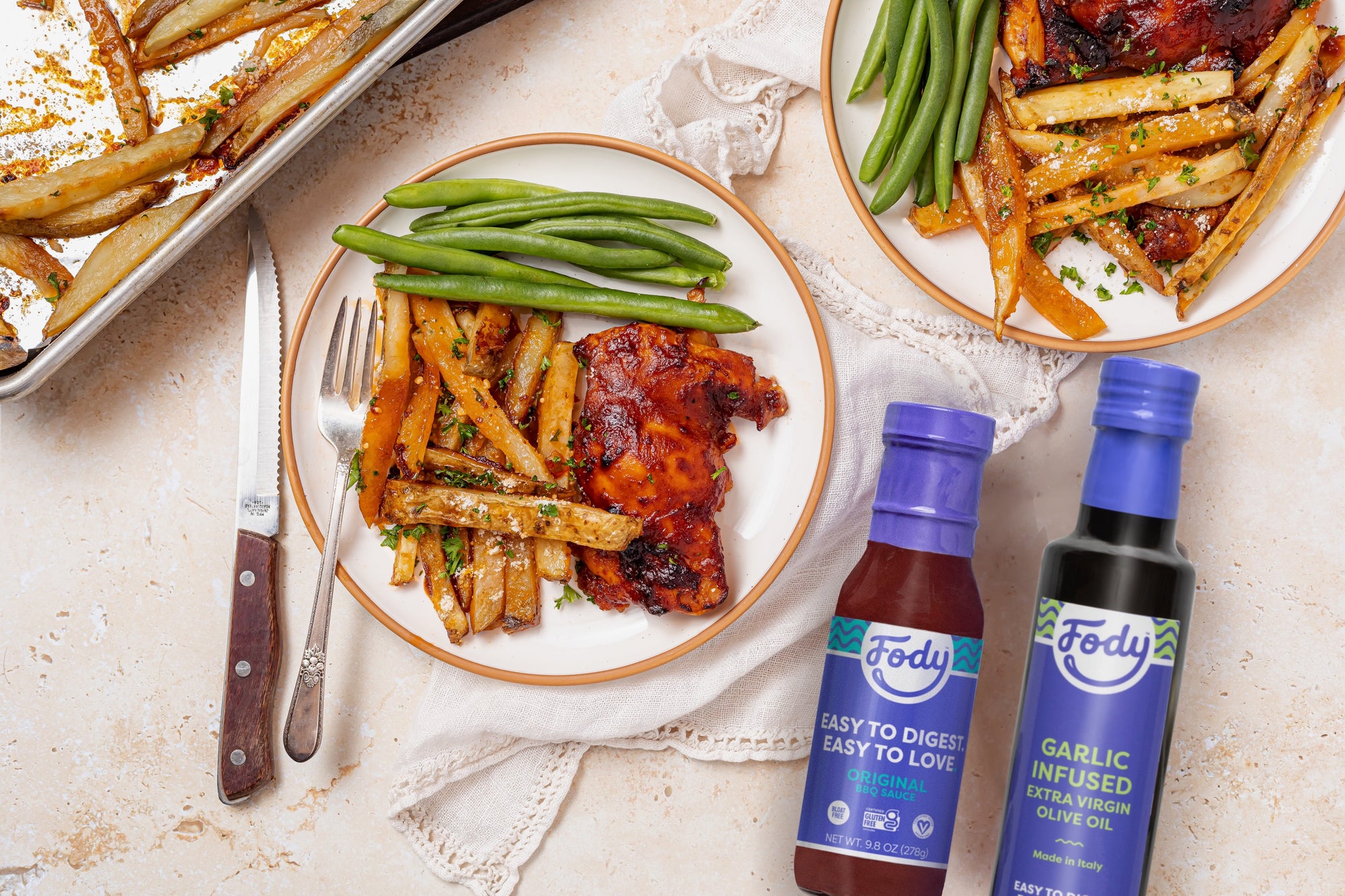 An image of Fody's Sheet Pan BBQ Chicken Thighs with Parmesan Truffle Fries laid out on white and brown plates, on a table next to two sauce bottles and elegant silver cutlery.