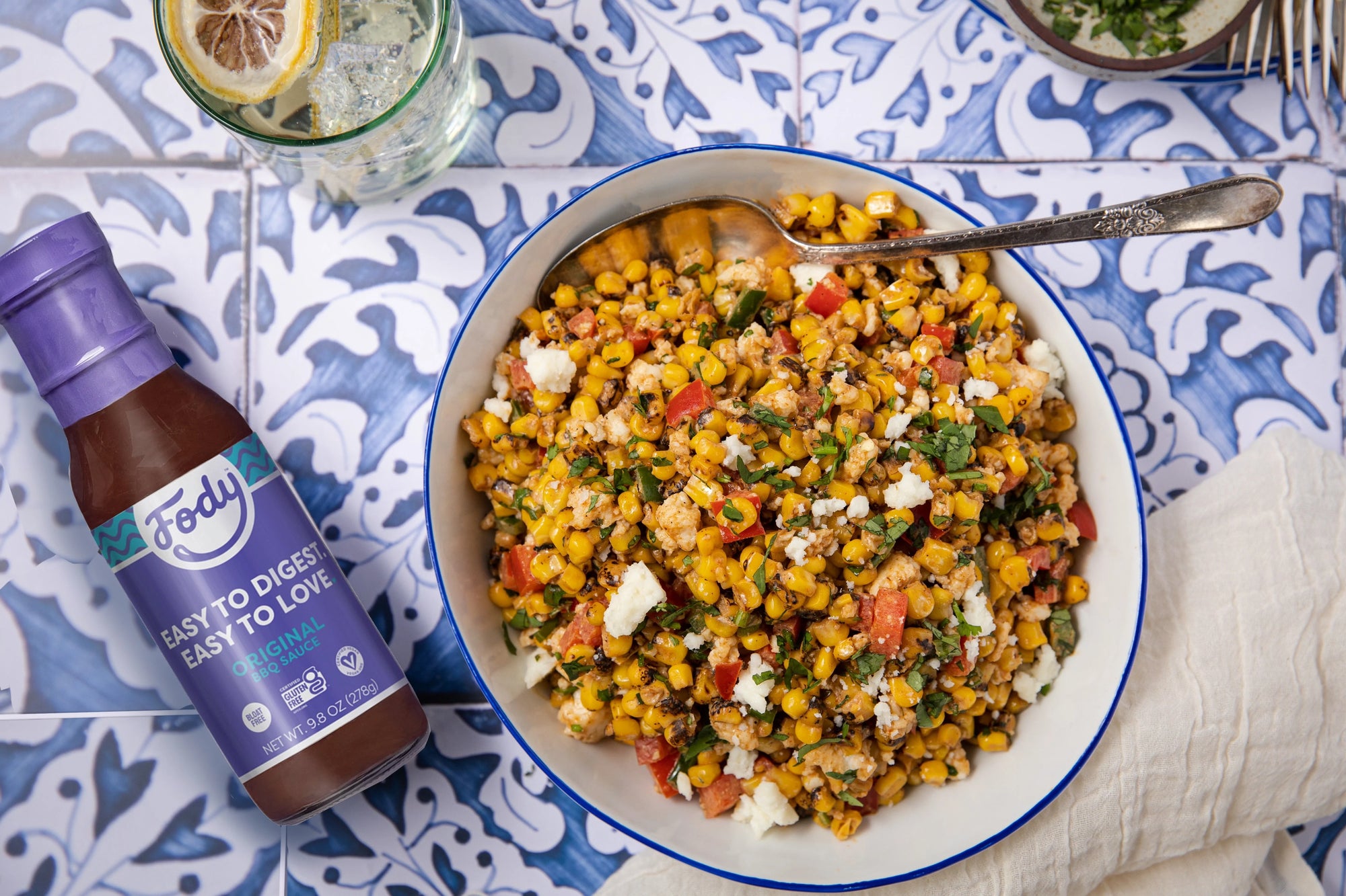 No bloat salad summer is just around the corner! Image: Fody’s BBQ corn salad in a white bowl against a backdrop of blue tiles beside a bottle of Fody’s Original BBQ Sauce.