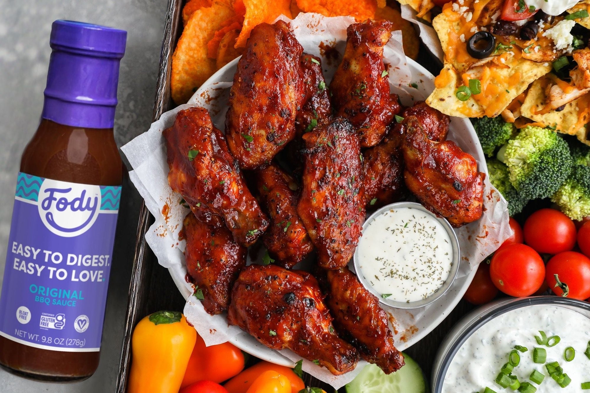 An image of Fody's baked honey BBQ chicken wings. Made with low FODMAP BBQ sauce, these chicken wings are on a plate beside dipping sauce, surrounded by vegetables.