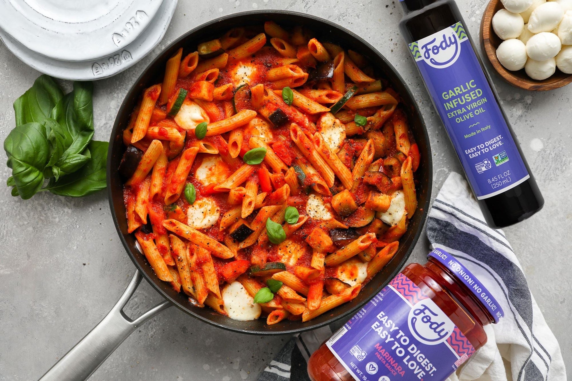An image of the perfect cheesy vegetarian pasta: Fody's Loaded Veggie Pasta with Mozzarella, tossed in red sauce, is displayed in a pan next to two jars of sauce, fresh mozzarella balls and fresh basil leaves.