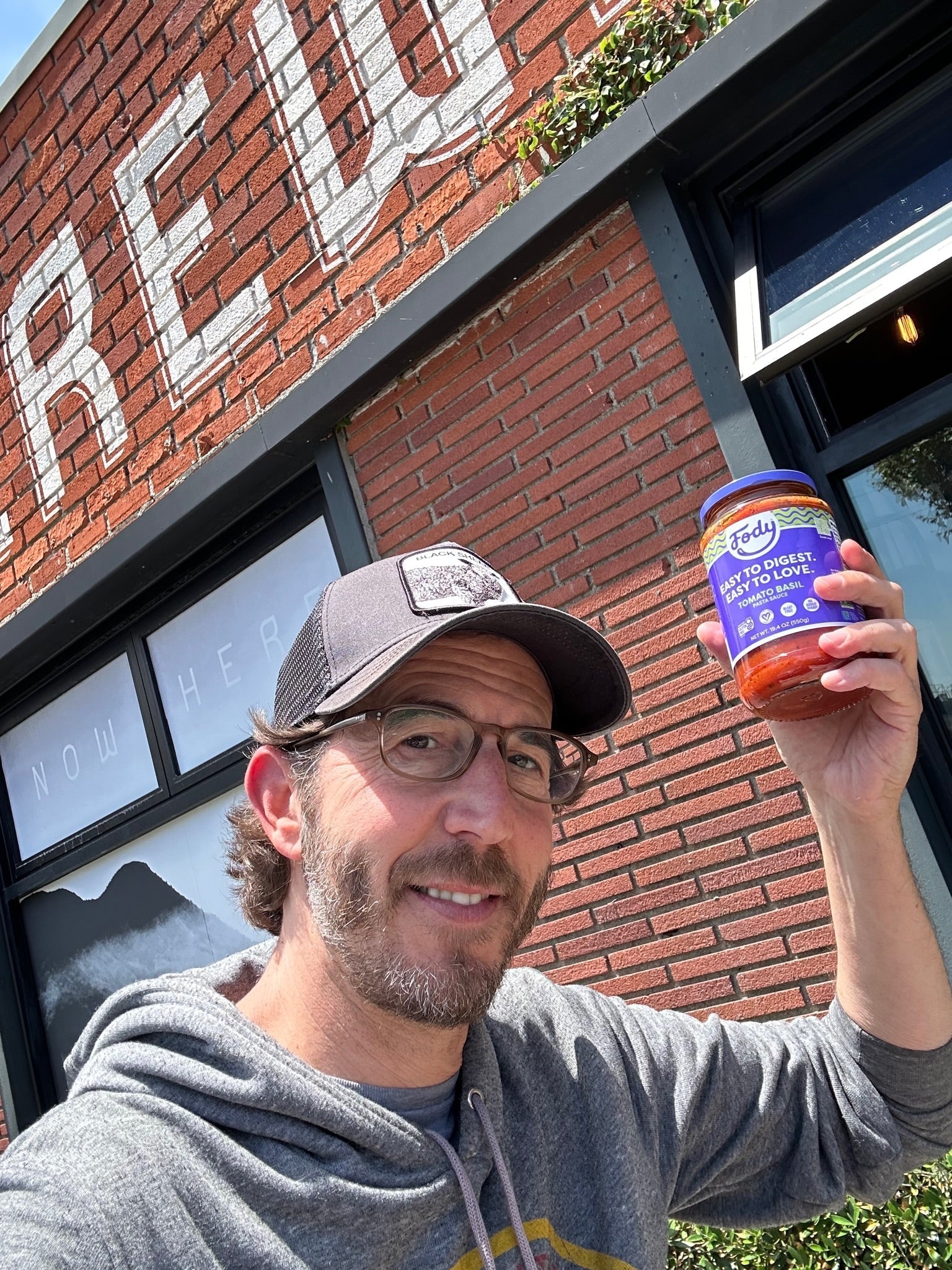 Man in glasses and a baseball cap holding a jar of Fody Tomato Basil pasta sauce outside a brick building with white painted signage.