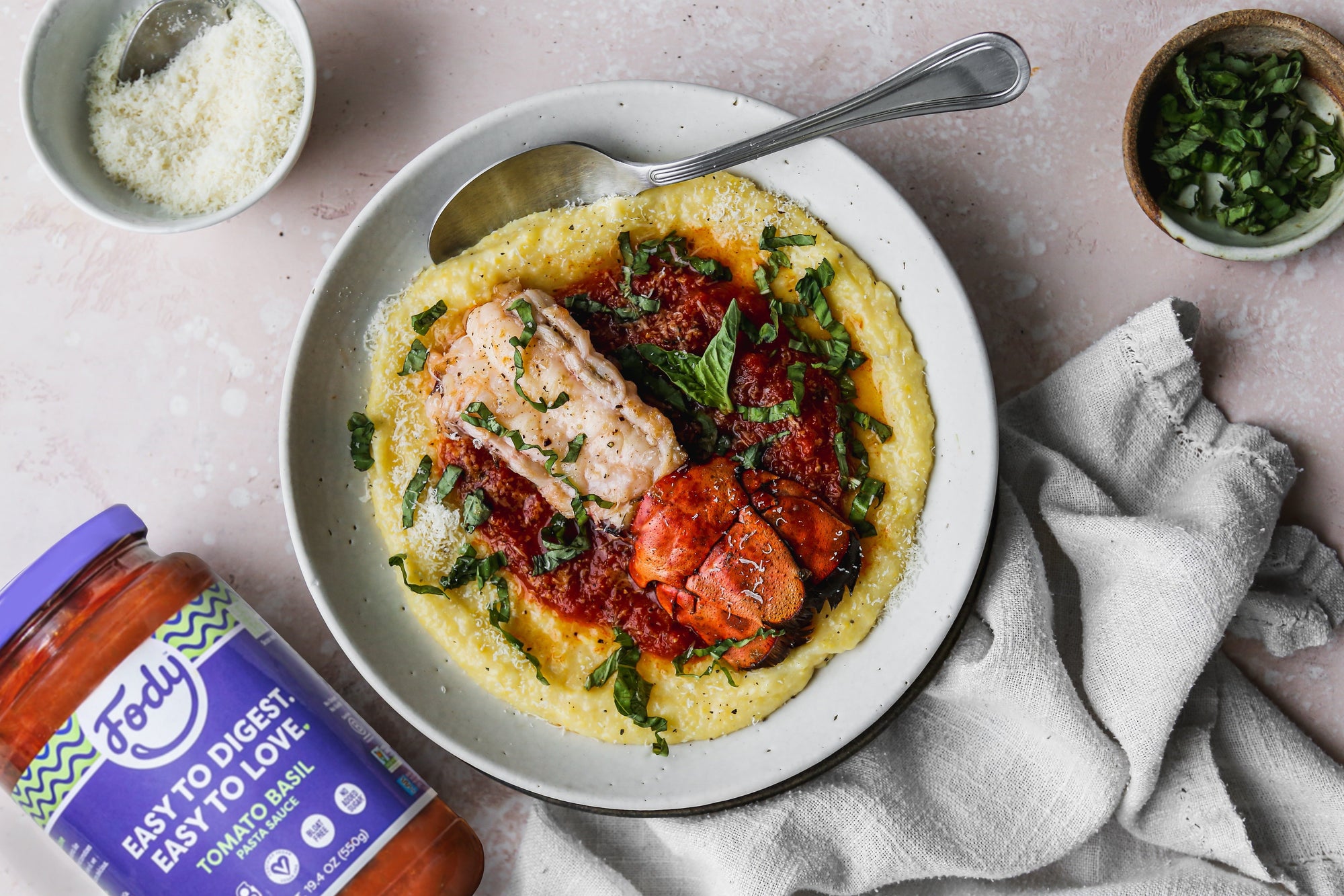 An image of Fody's broiled lobster with creamy tomato polenta. The lobster and polenta dish is sitting on a table in a bowl beside a jar of Fody's marinara sauce.