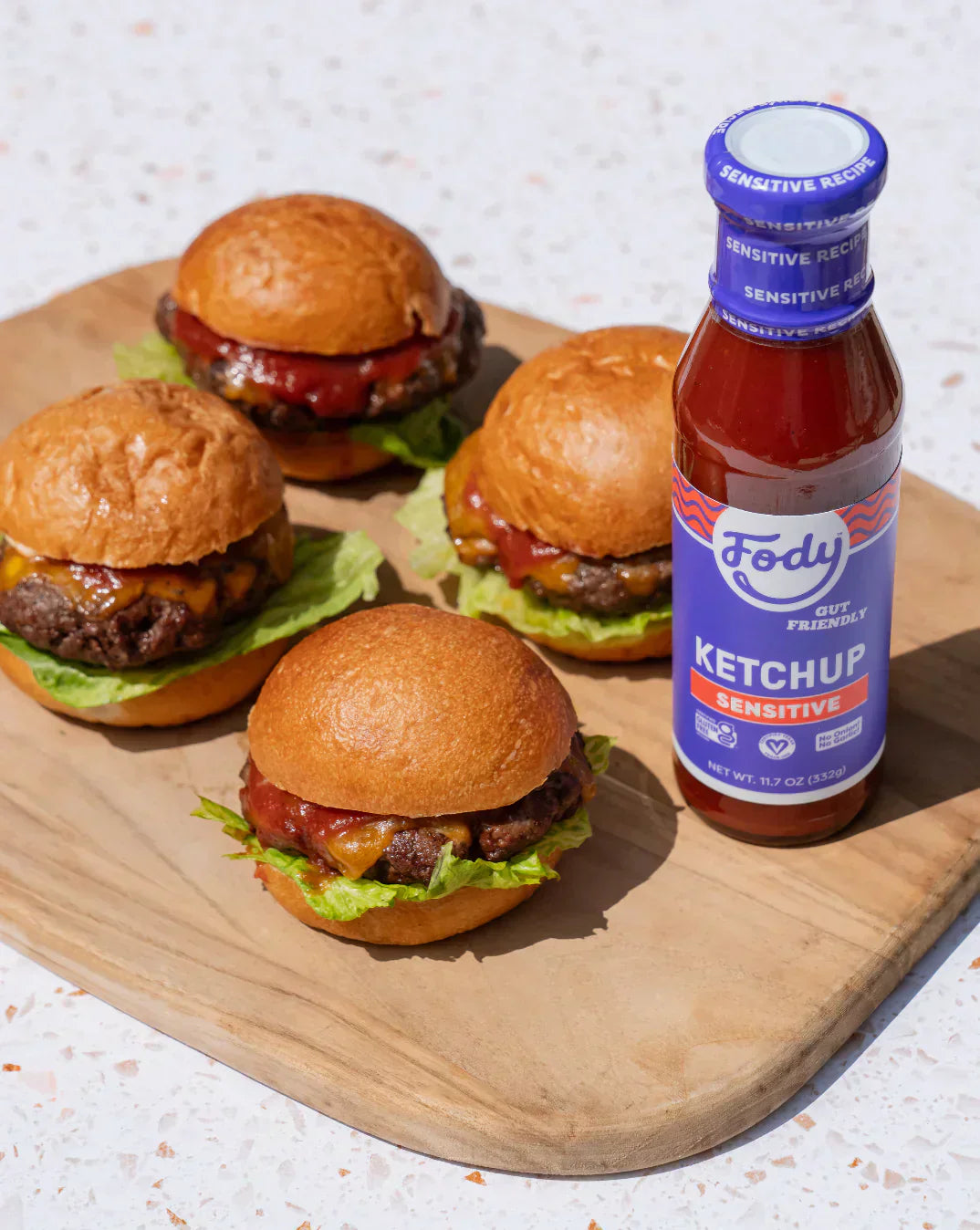 Fody Low FODMAP ketchup bottle beside cheeseburgers on a wooden board, showing an IBS-friendly condiment option made without onion or garlic.