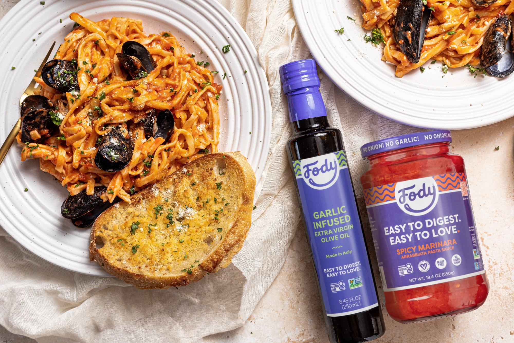 An image of Fody’s Spicy Mussels and Pasta with Garlic Bread. This low FODMAP seafood pasta is on a white plate beside a piece of garlic bread. A jar of FODY's olive oil and spicy marinara are also in the frame.