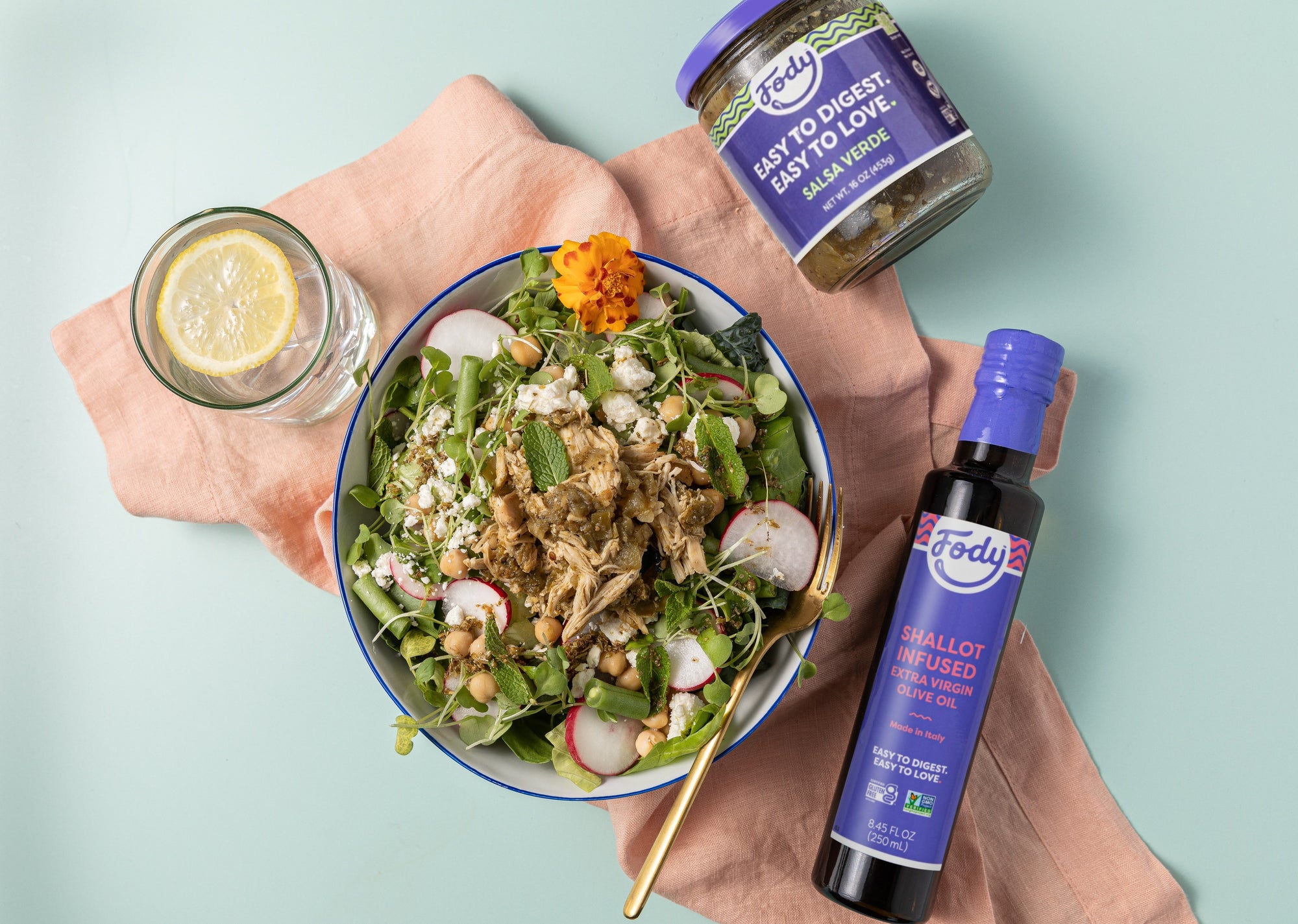 An image of Fody’s Spring Greens Salad with Slow Cooker Salsa Verde Chicken in a bowl on a light turquoise table beside bottles of Fody's olive oil and Fody's salsa verde.