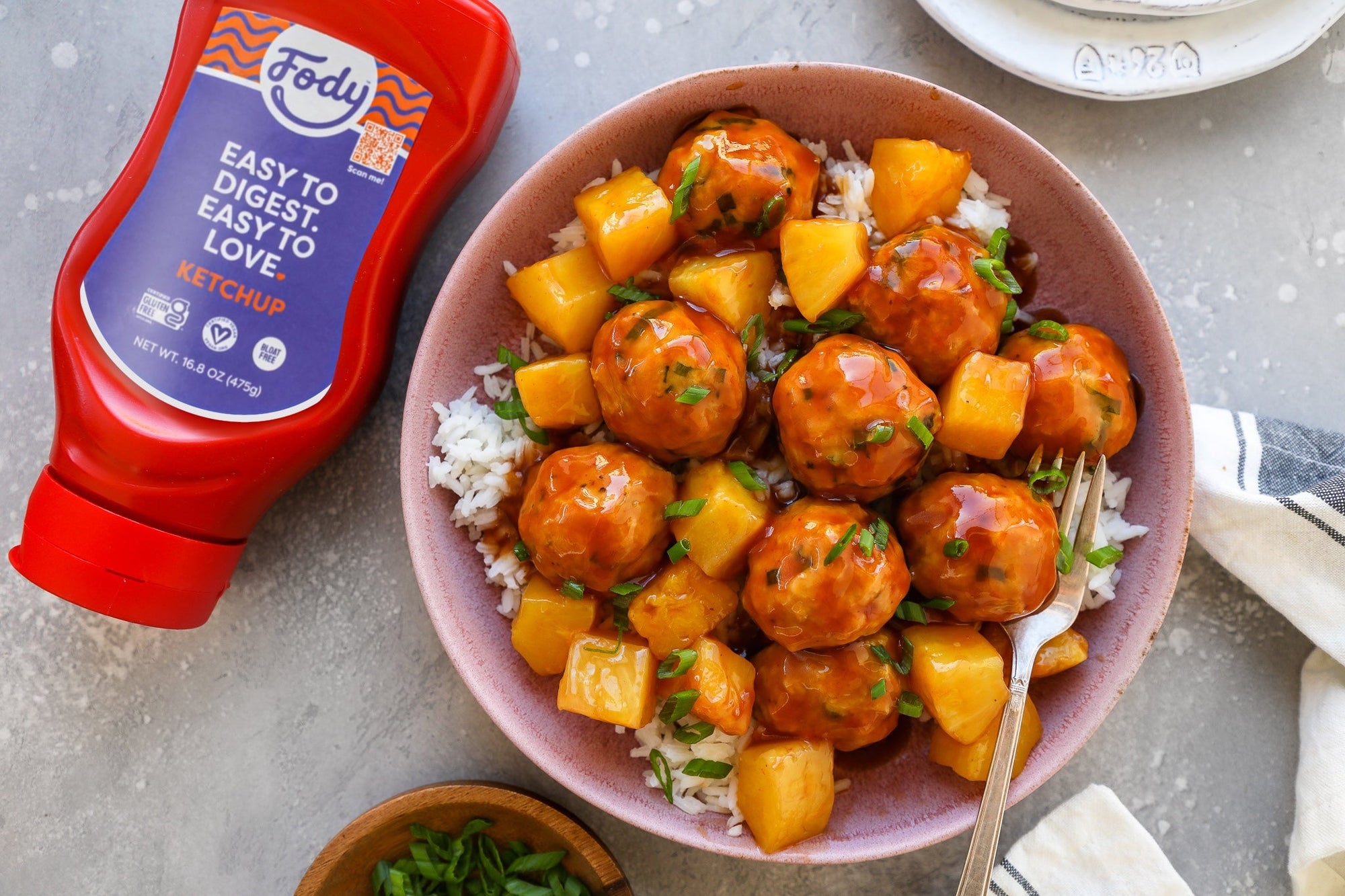 A bowl of sweet and sour chicken meatballs over rice, beside a bottle of Fody's no onion no garlic ketchup