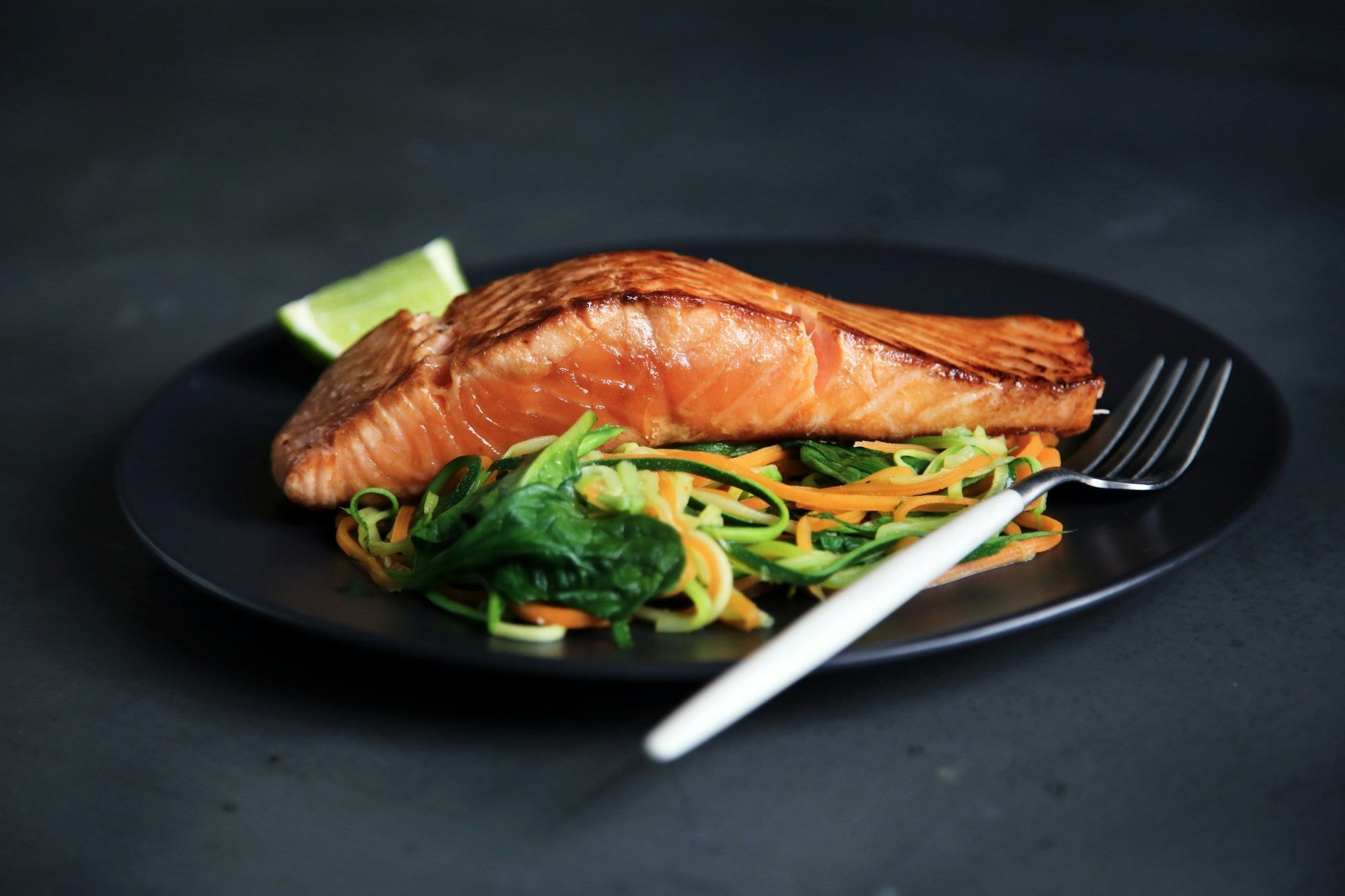 A plate of grilled fish with cooked vegetables and a fork on a dark surface, illustrating safe Low FODMAP protein and vegetable options for people managing IBS.