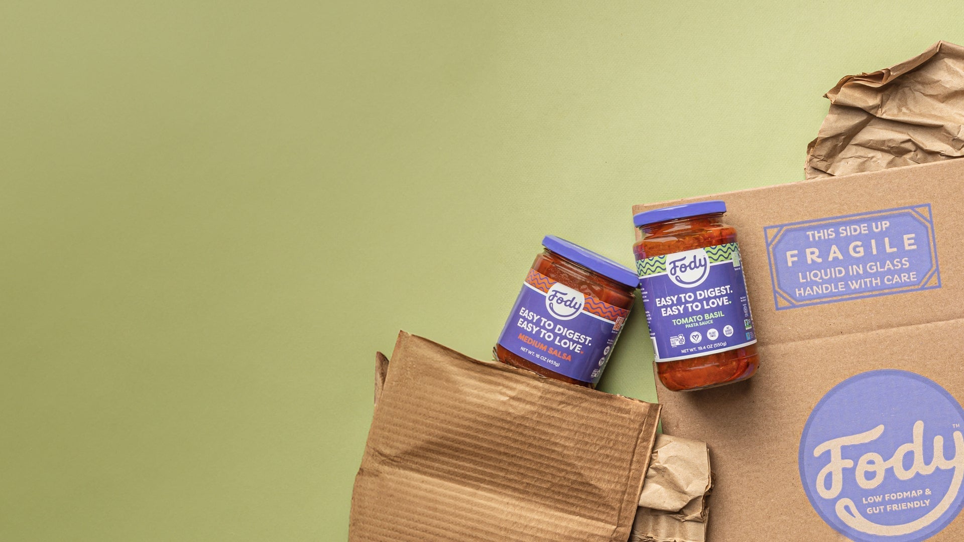 Two jars of tomato-based sauce are placed beside crumpled paper and a Fody-branded shipping box on a light green background.
