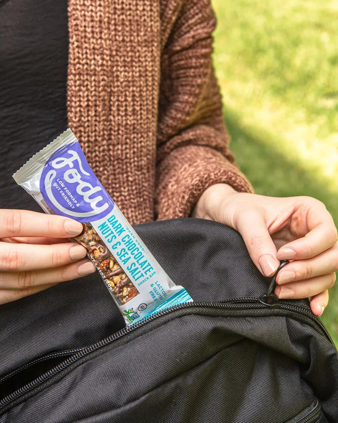 Fody Dark Chocolate Nuts and Sea Salt snack bar held in hand while opening a black backpack outdoors