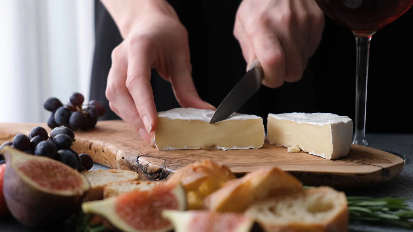 Cheese and gut health go together like cheese and well…everything! Image: A pair of hands cutting a wedge of camembert on a cutting board sprinkled with figs and sliced baguette.