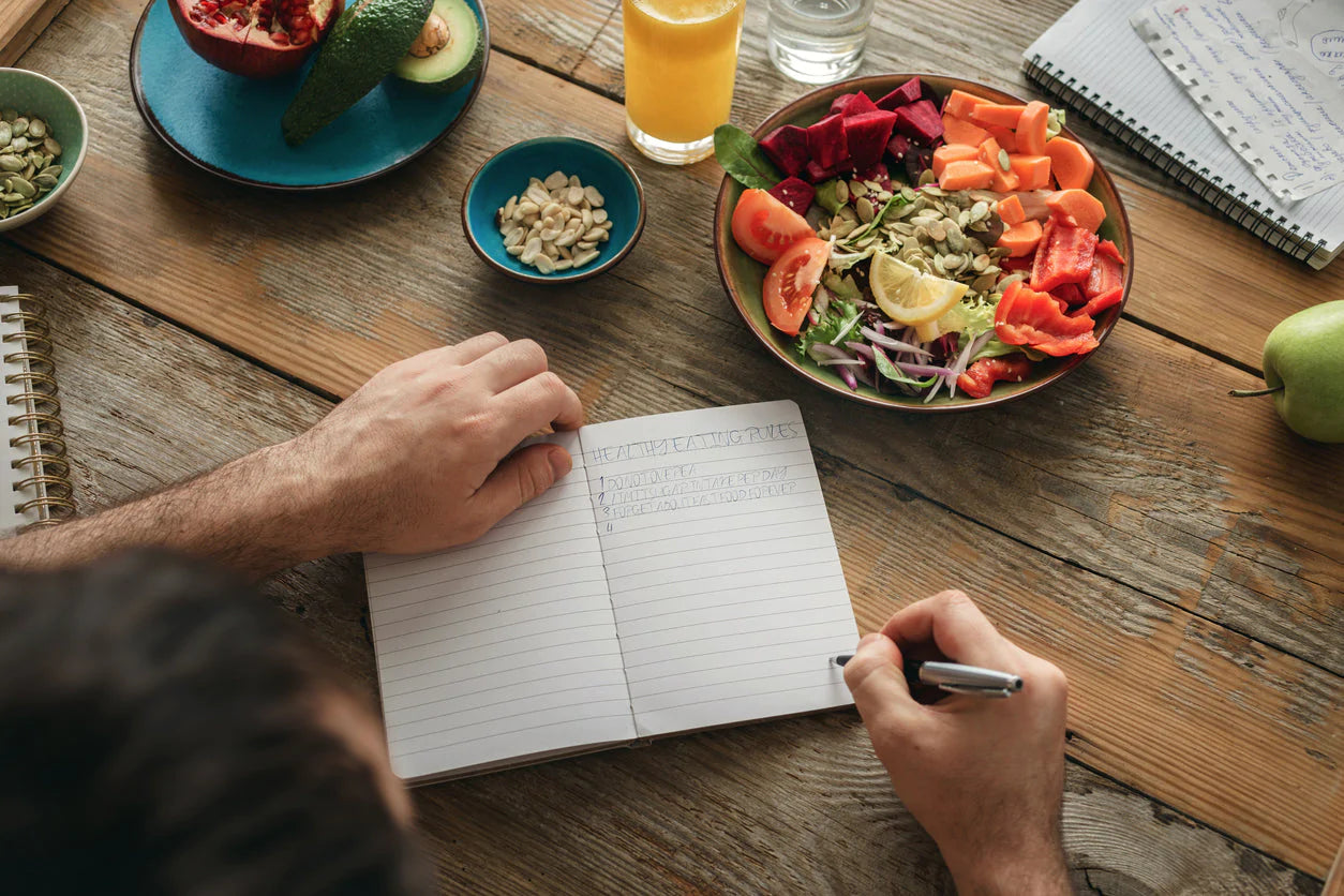 A man is writing in a food journal with a salad and a bowl of nuts on the table beside him. He is keeping a food journal to help identify the triggers of his sensitive stomach.