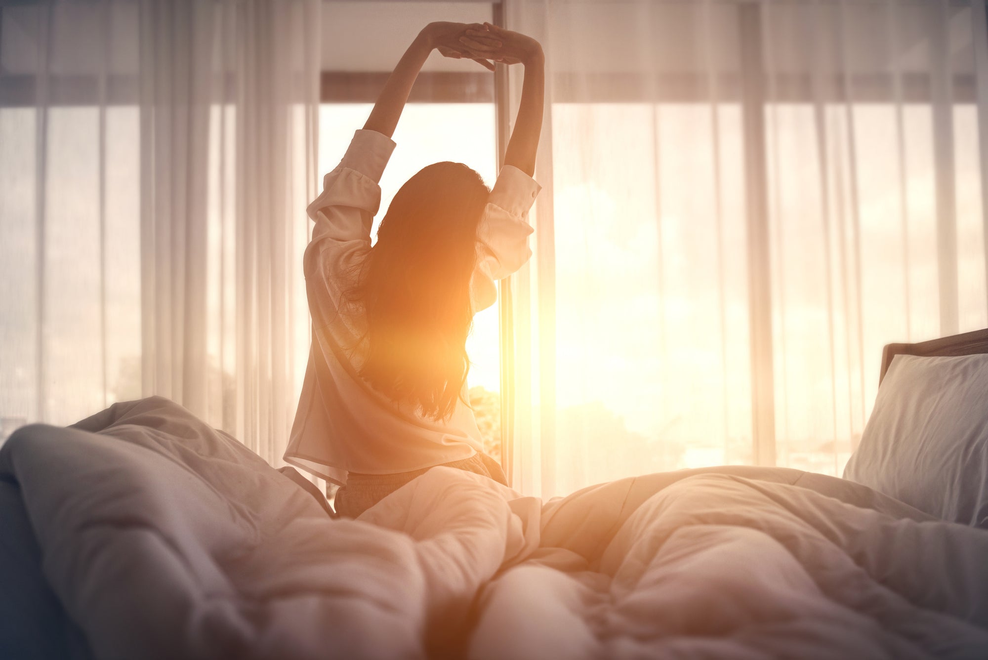 An image of a woman sitting and stretching on her bed against a bright sunrise.
