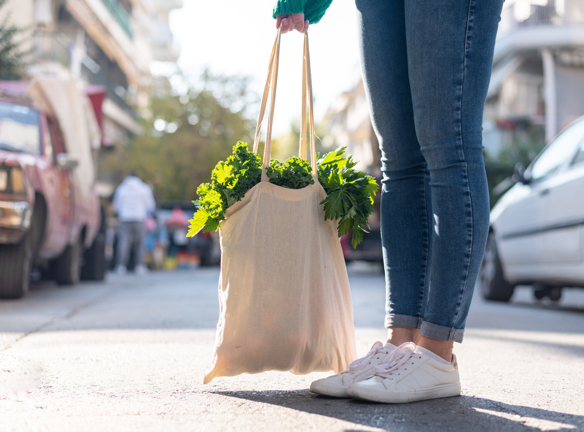 An image of a woman’s feet and legs standing on a sidewalk. She is wearing jeans and white sneakers, and carrying a reusable shopping bag filled with leafy greens to help her pursue plant-based gut health.