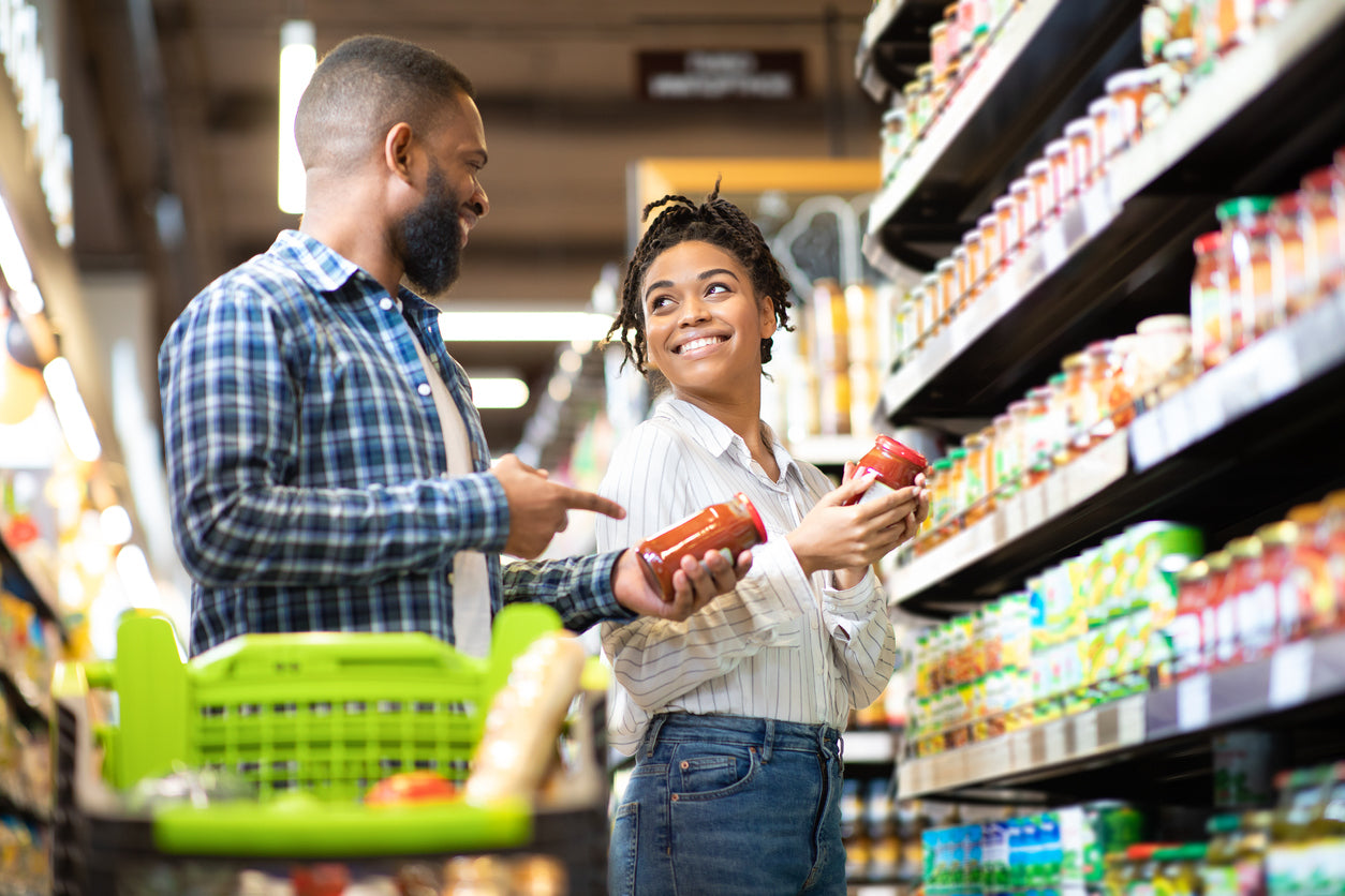 Gut healthy eating on a budget can be simple! Image: A young couple grocery shopping and comparing two bottles of red sauce.