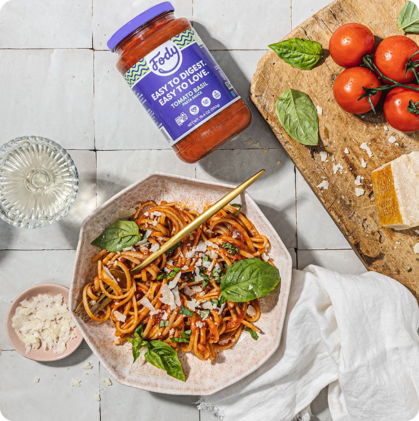 Overhead view of a plate of pasta with red sauce beside a jar of Fody pasta sauce and fresh tomatoes, an easy Low FODMAP dinner idea