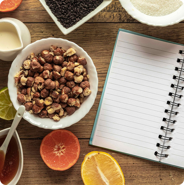 Blank notepad and pen on a wooden table beside halved oranges, berries, nuts, and fresh vegetables, set up for planning midweek dinners
