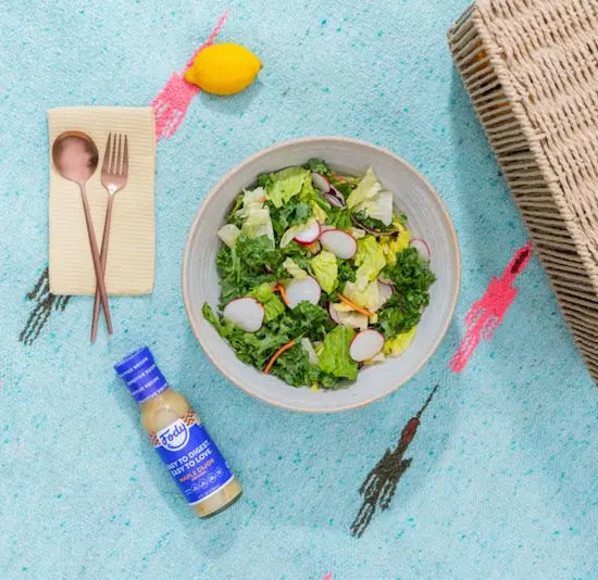 Bowl of green salad with kale, lettuce, sliced radishes and shredded carrot beside a bottle of Fody salad dressing on a turquoise cloth