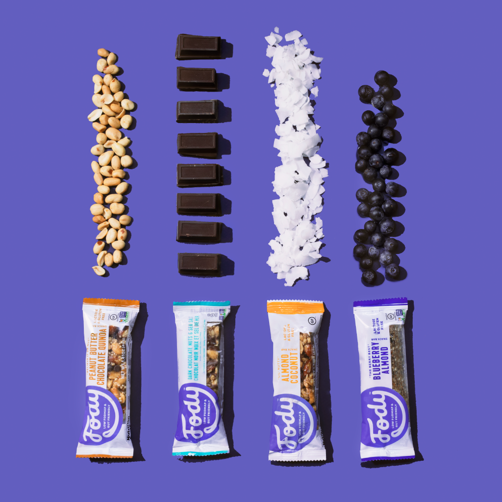 Four Fody snack bars on purple, each below a row of a key ingredient: peanuts, dark chocolate squares, coconut flakes and fresh blueberries