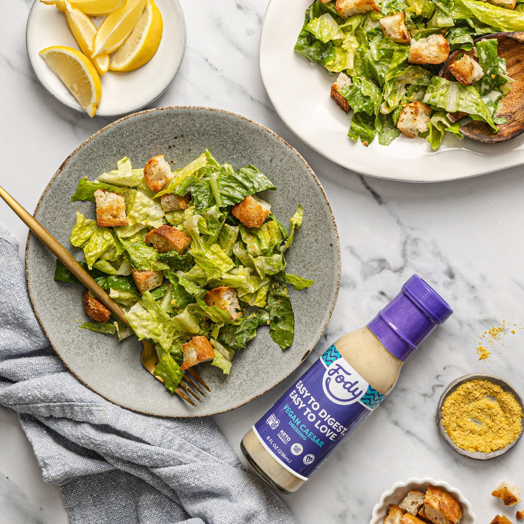 Romaine salad with golden croutons on a grey plate beside a bottle of Fody Vegan Caesar Dressing, with extra salad in a serving dish behind