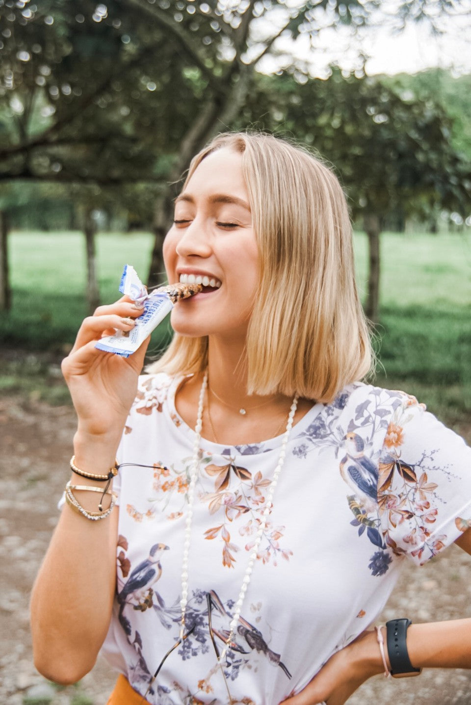Smiling woman takes a bite of a Fody snack bar outdoors under the trees, an individually wrapped Low FODMAP snack that is easy to carry